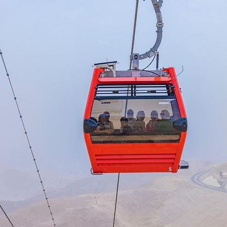 منتجع توليب الجلالة ماجستيك عين السخنة المظهر الخارجي الصورة The photo shows a red gondola lift suspended in the air, traveling along cables. Inside the gondola, a group of people can be seen. The background features a hazy landscape, possibly mountains or hills, indicating an elevated area. The scene conveys 