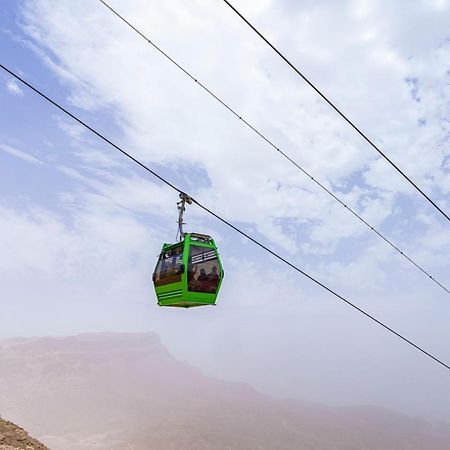 منتجع توليب الجلالة ماجستيك عين السخنة المظهر الخارجي الصورة The photo shows a green cable car suspended in the air, traveling along overhead cables. The backdrop features a hazy landscape, likely mountainous or elevated terrain, with a cloudy sky. The scene suggests a scenic transportation method, possibly us