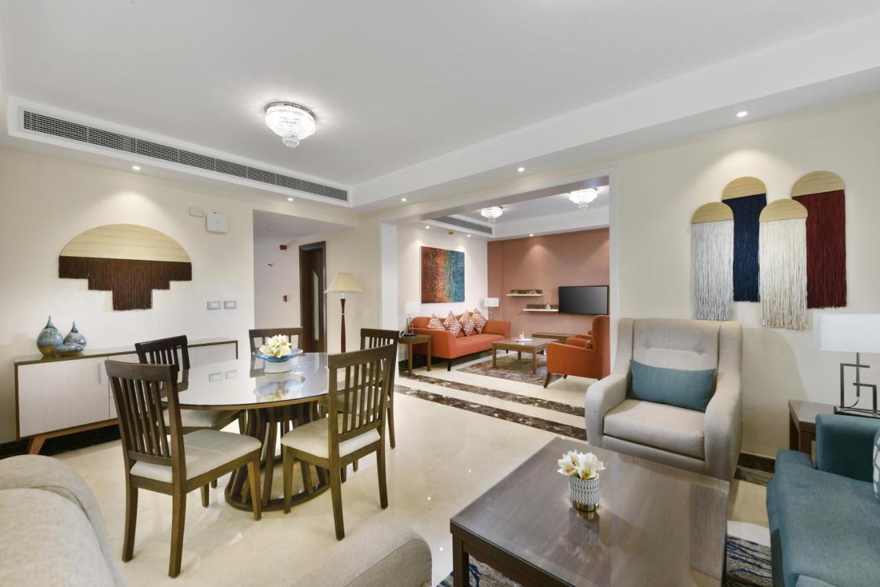 منتجع توليب الجلالة ماجستيك عين السخنة المظهر الخارجي الصورة The photo shows a modern living room space that is elegantly designed. It features a seating area with a comfortable sofa and armchairs, complemented by a decorative coffee table. There is a dining table with several wooden chairs, providing a cozy a