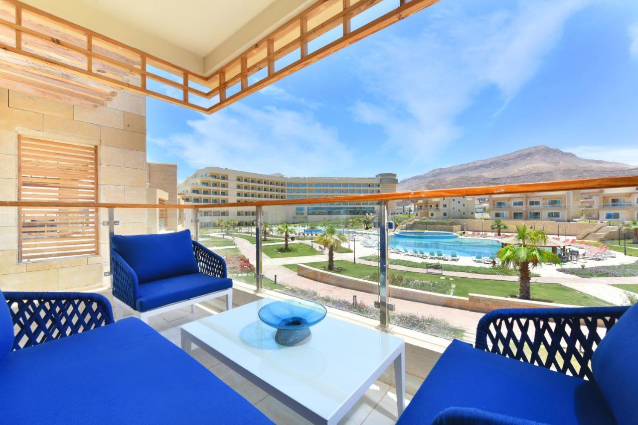 منتجع توليب الجلالة ماجستيك عين السخنة المظهر الخارجي الصورة The photo showcases a balcony view featuring two blue chairs and a table with a decorative bowl. The balcony overlooks a landscape that includes a swimming pool and manicured lawns. In the background, there are multiple buildings and palm trees, all 