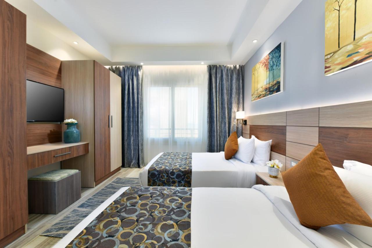 منتجع توليب الجلالة ماجستيك عين السخنة المظهر الخارجي الصورة The photo shows a well-decorated hotel room with two twin beds, each adorned with white linens and brown decorative pillows. There's a modern wooden wardrobe in the corner, alongside a flat-screen TV mounted on the wall. The room features large windo