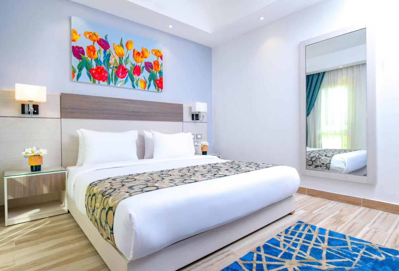 منتجع توليب الجلالة ماجستيك عين السخنة المظهر الخارجي الصورة The photo shows a modern hotel bedroom. It features a large bed with white linens and a decorative throw blanket. The room has light wooden flooring and is bright, with ample natural light coming from a window. There is a colorful painting of flowers