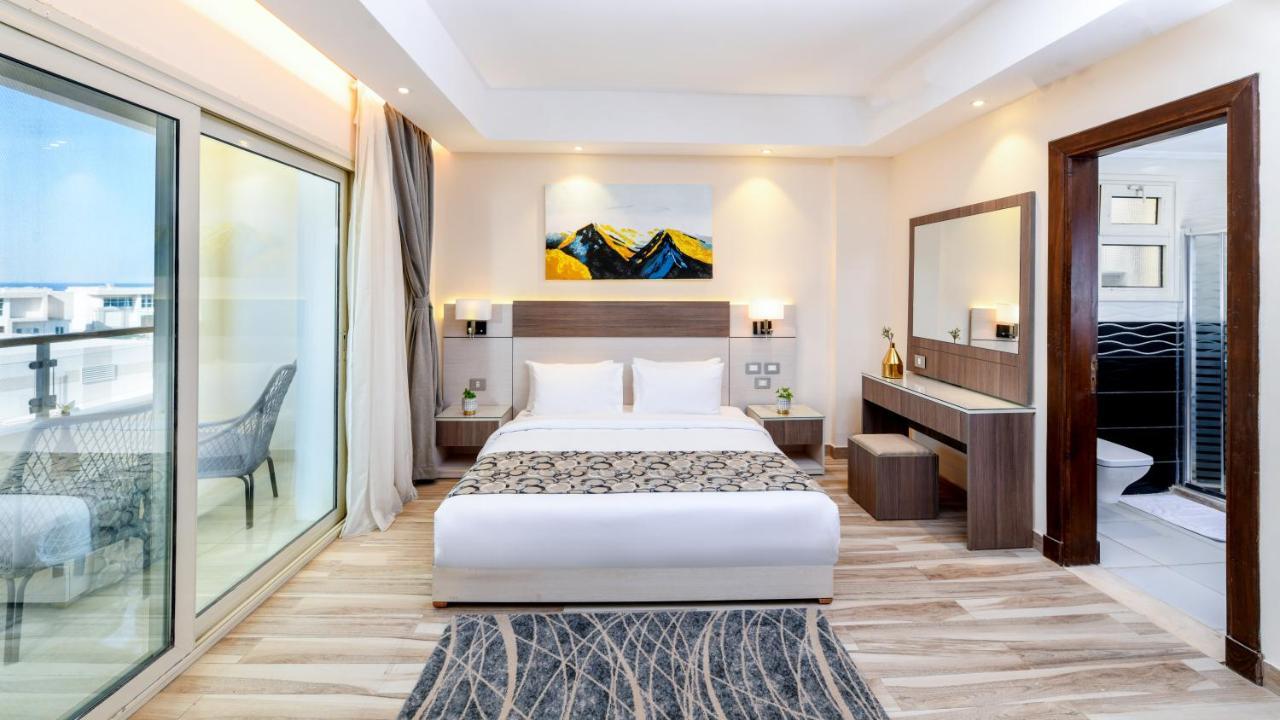 منتجع توليب الجلالة ماجستيك عين السخنة المظهر الخارجي الصورة The photo shows a modern hotel room featuring a large bed with white linens and a decorative throw. The room has a neutral color palette, accented with warm lighting. There is a piece of artwork above the bed depicting mountains. To the left, there a