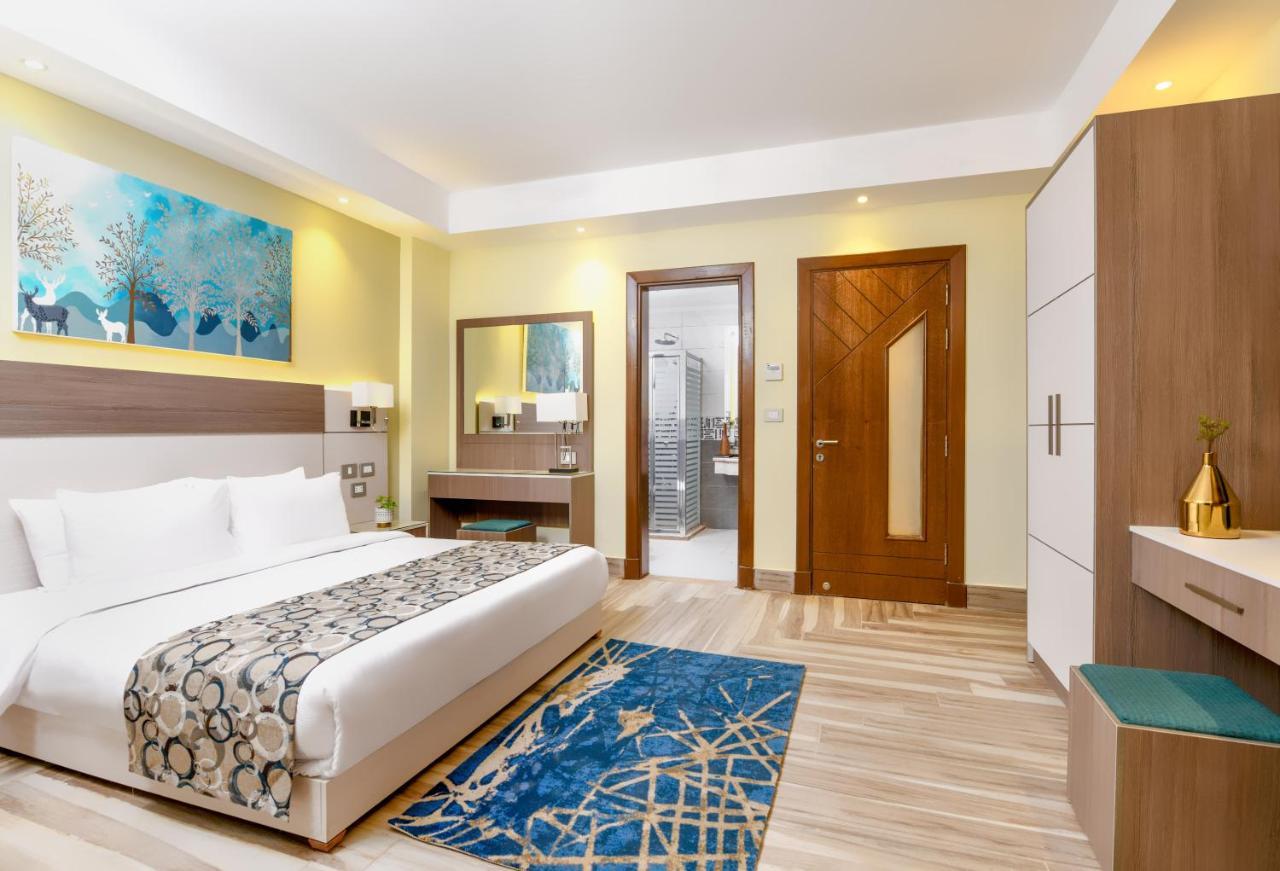 منتجع توليب الجلالة ماجستيك عين السخنة المظهر الخارجي الصورة The photo shows a well-decorated hotel room. There is a large bed with white linens and a decorative throw blanket. The room features a wooden wardrobe and a bedside table with lamps. A colorful rug is placed on the floor, adding warmth to the space.