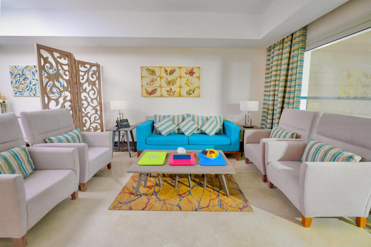 منتجع توليب الجلالة ماجستيك عين السخنة المظهر الخارجي الصورة The photo shows a stylish and colorful living room. It features a bright blue sofa with patterned cushions, complemented by two light gray armchairs. In front of the sofa, there's a wooden coffee table with vibrant trays in pink, green, and yellow. A