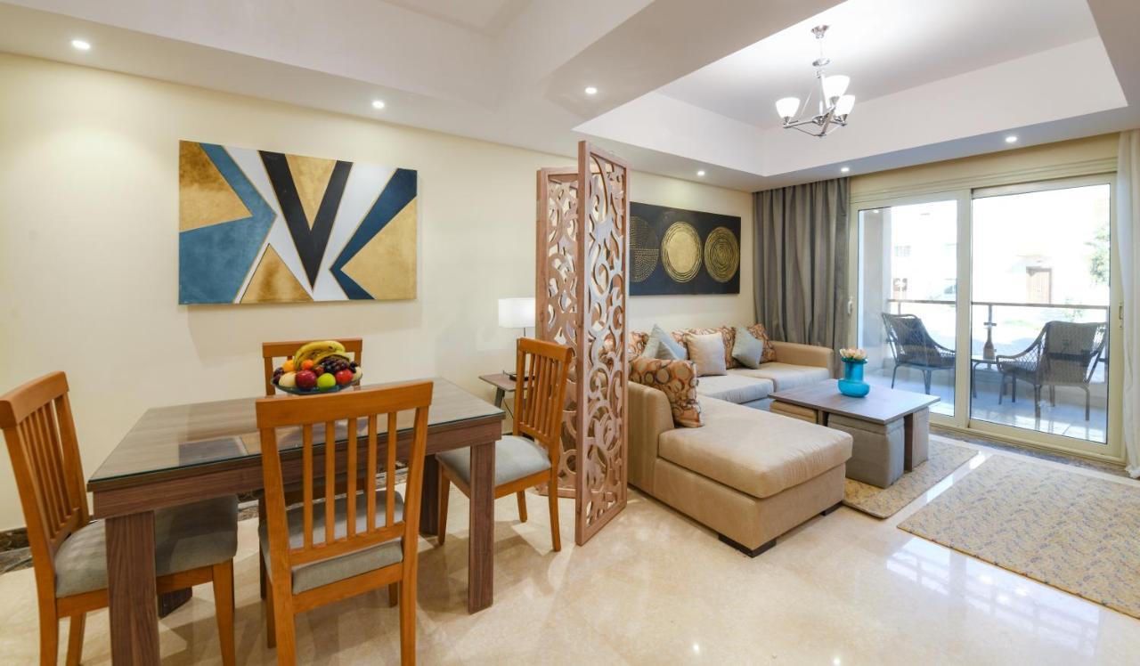 منتجع توليب الجلالة ماجستيك عين السخنة المظهر الخارجي الصورة The photo shows a modern and stylish interior of a living space. It features a seating area with a plush sofa and decorative cushions, complemented by a wooden coffee table. There is a dining table with wooden chairs nearby. 

On the walls, there are