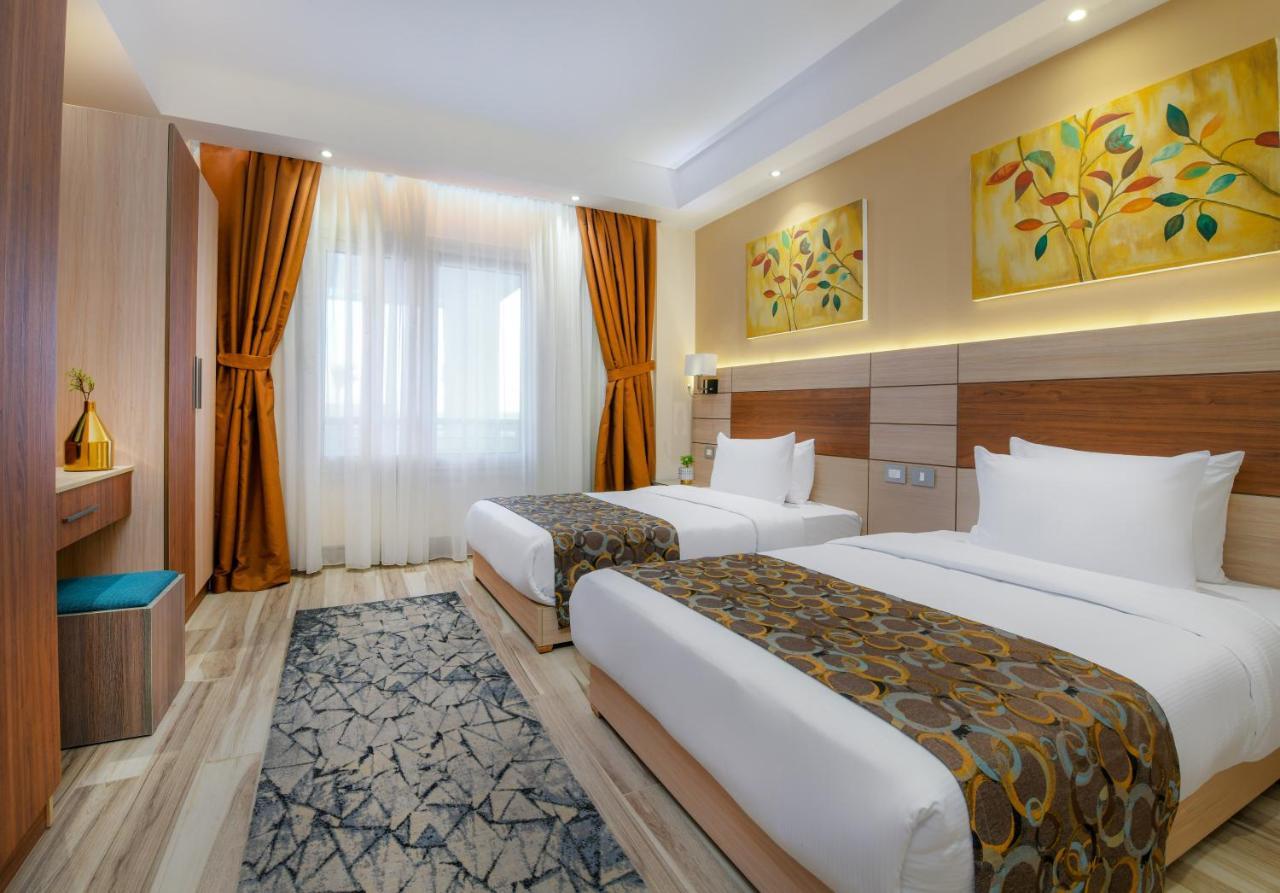 منتجع توليب الجلالة ماجستيك عين السخنة المظهر الخارجي الصورة The photo shows a modern hotel room with two single beds, each covered with white linens and decorative brown bed throws. The walls are painted in warm colors, and there are two pieces of artwork featuring floral and leaf designs above the beds. The 