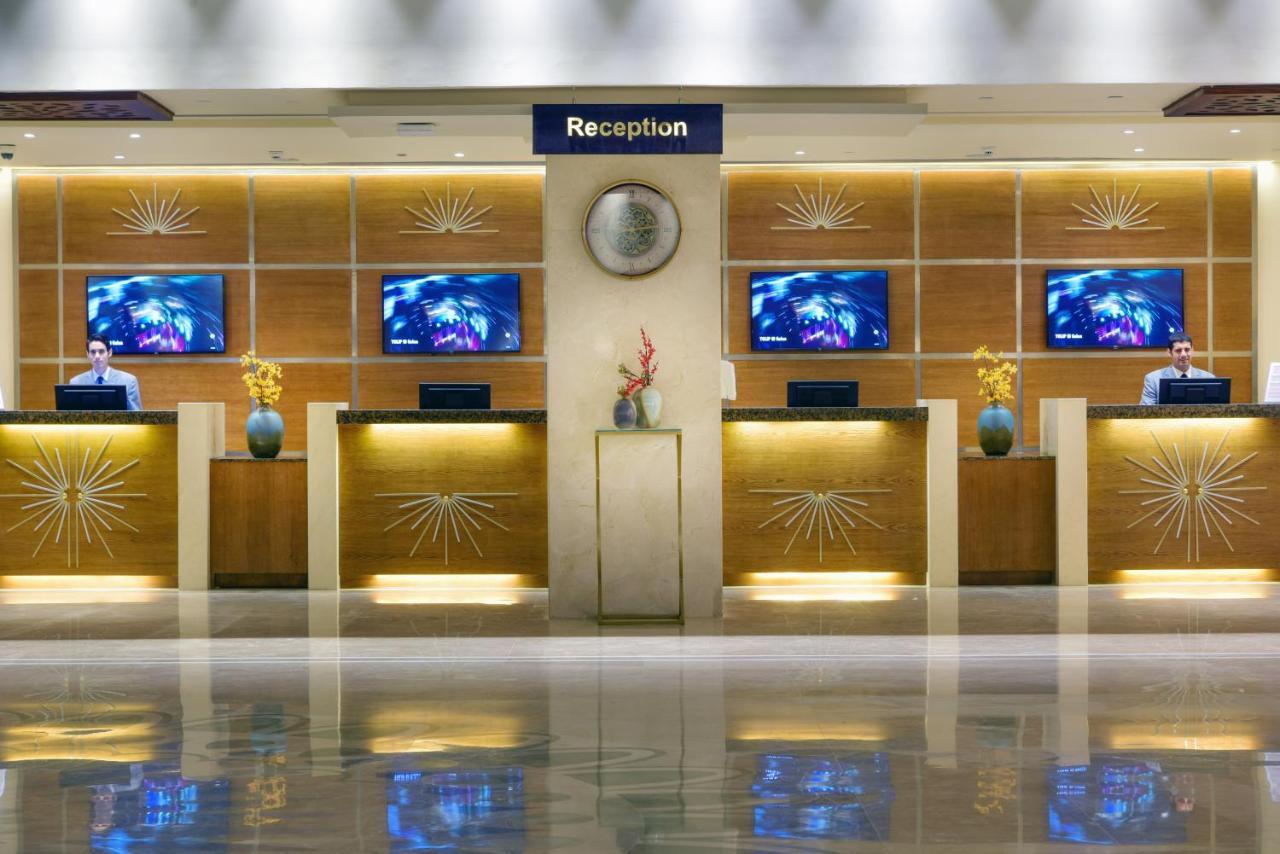 منتجع توليب الجلالة ماجستيك عين السخنة المظهر الخارجي الصورة The image shows a hotel reception area. There are several reception desks lined up against a wall with decorative paneling. Each desk has a staff member, and above the desks, there are screens displaying information. A clock is visible on the wall, a
