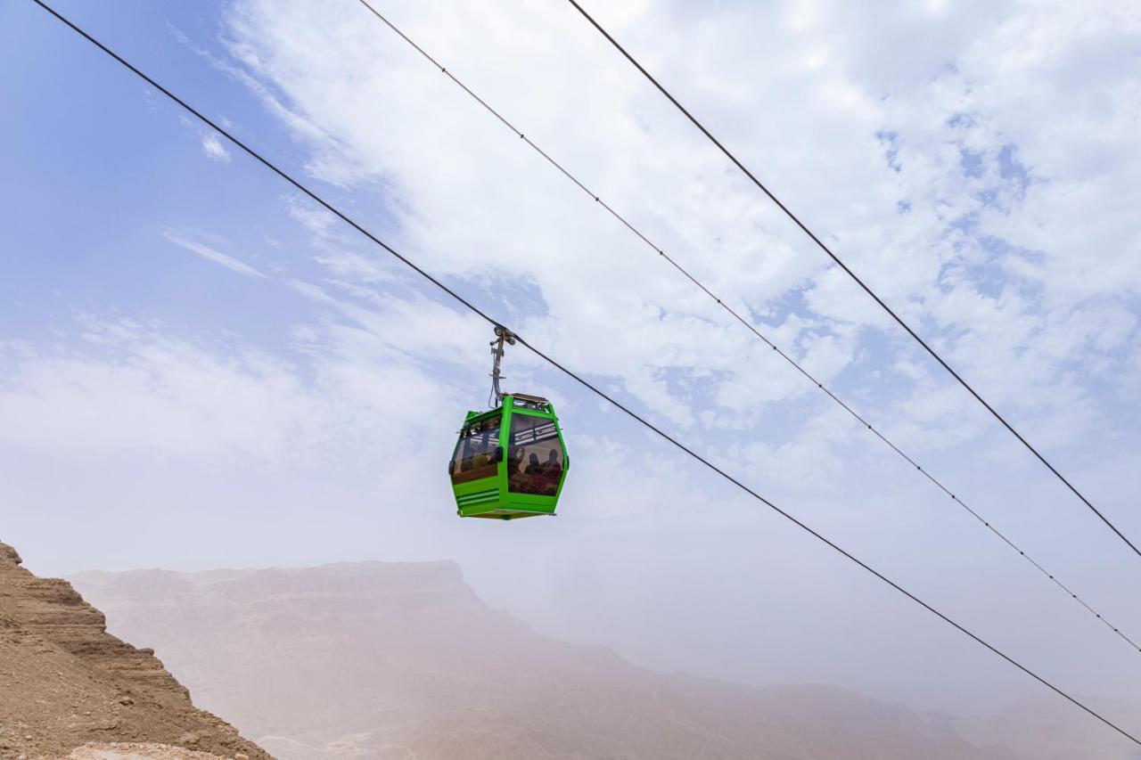 منتجع توليب الجلالة ماجستيك عين السخنة المظهر الخارجي الصورة The photo shows a green cable car suspended in the air, traveling along overhead cables. The backdrop features a hazy landscape, likely mountainous or elevated terrain, with a cloudy sky. The scene suggests a scenic transportation method, possibly us