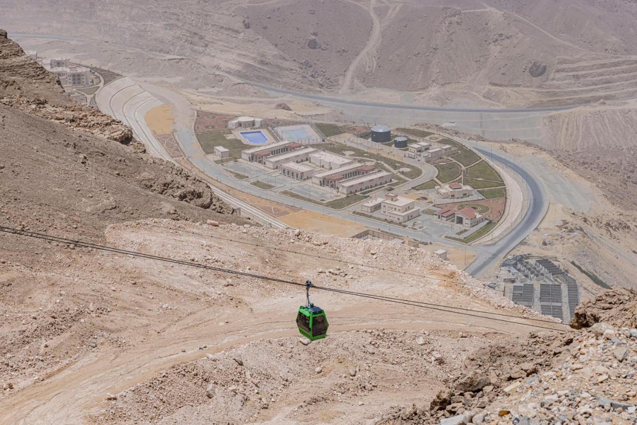 منتجع توليب الجلالة ماجستيك عين السخنة المظهر الخارجي الصورة The photo shows a mountainous landscape with a steep, dry terrain. In the foreground, there is a green cable car or gondola traveling along a cable. Below, a complex of buildings and roads can be seen, likely part of a resort or recreational area. Th