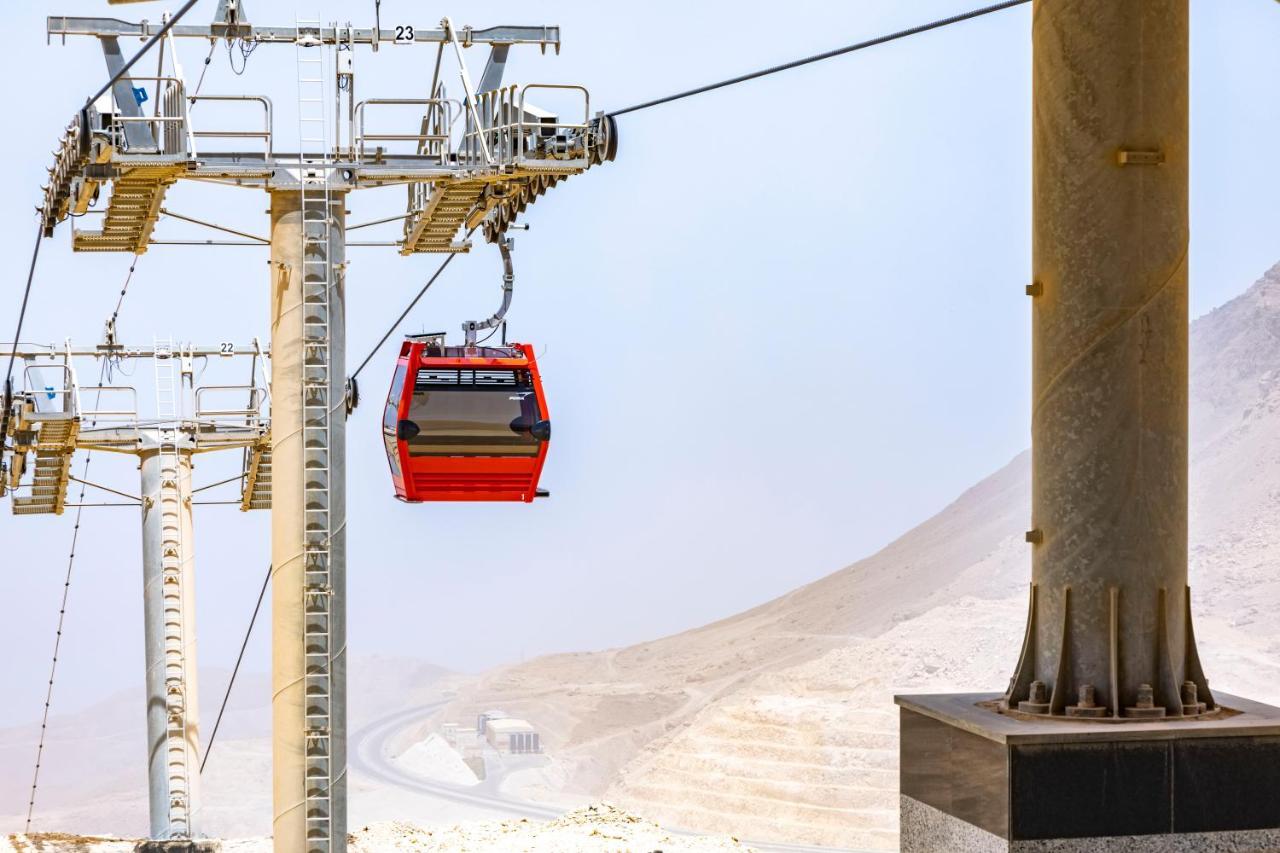 منتجع توليب الجلالة ماجستيك عين السخنة المظهر الخارجي الصورة The photo shows a cable car system with a single red gondola suspended mid-air. It appears to be part of a larger cable car infrastructure, with metal pylons supporting the cables above. The background features a hazy landscape, likely characterized 