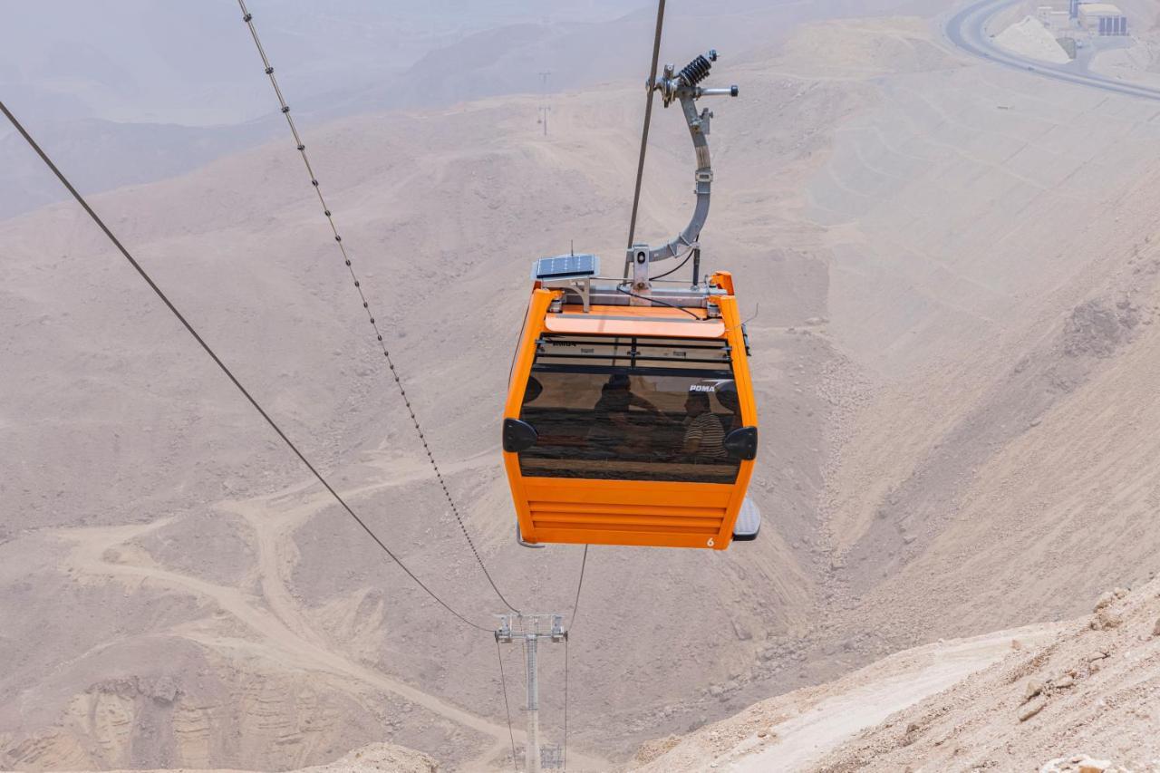 منتجع توليب الجلالة ماجستيك عين السخنة المظهر الخارجي الصورة The photo shows an orange cable car suspended in the air, traveling along a set of cables. It appears to be located in a mountainous or hilly area, with rugged terrain and dry, rocky landscapes visible below. The background features slopes that seem 