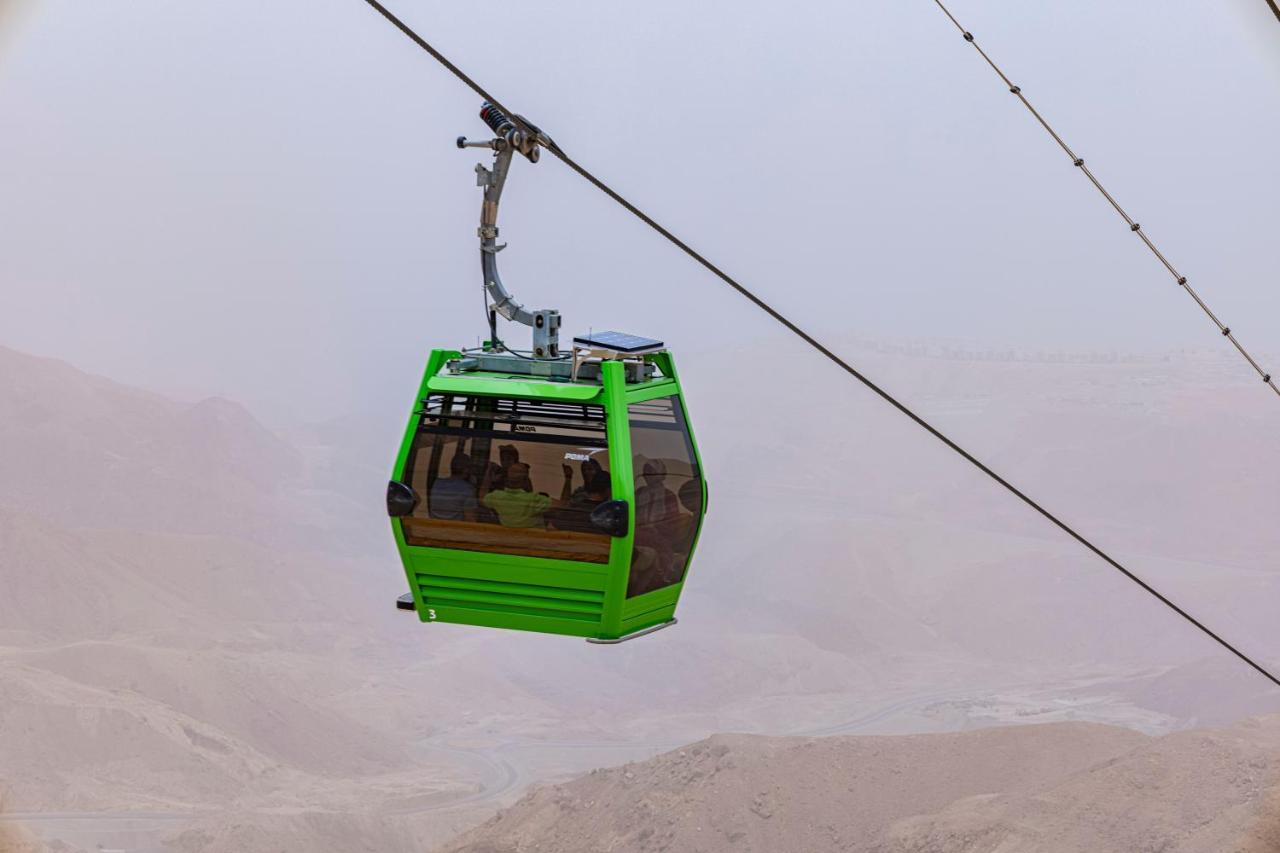 منتجع توليب الجلالة ماجستيك عين السخنة المظهر الخارجي الصورة The photo shows a green cable car suspended in the air, moving along a wire. The cable car appears to be traversing a mountainous landscape, which looks rocky and arid. The surrounding environment is somewhat foggy or hazy, suggesting limited visibil