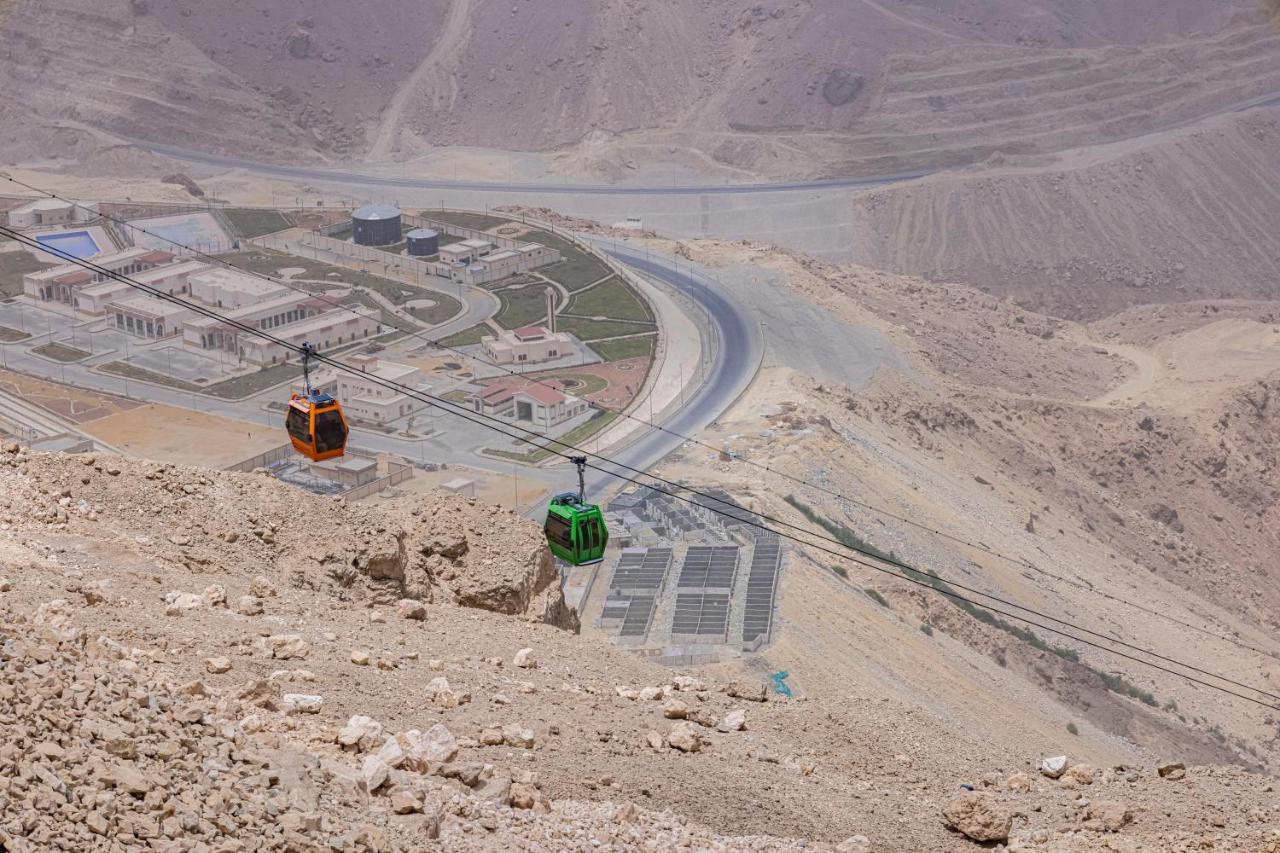 منتجع توليب الجلالة ماجستيك عين السخنة المظهر الخارجي الصورة The photo shows a scenic view from a high elevation, featuring a cable car system. Two cable cars are visible, one orange and one green, traveling along cables down the mountainside. Below, there appears to be a developed area with roads and light st