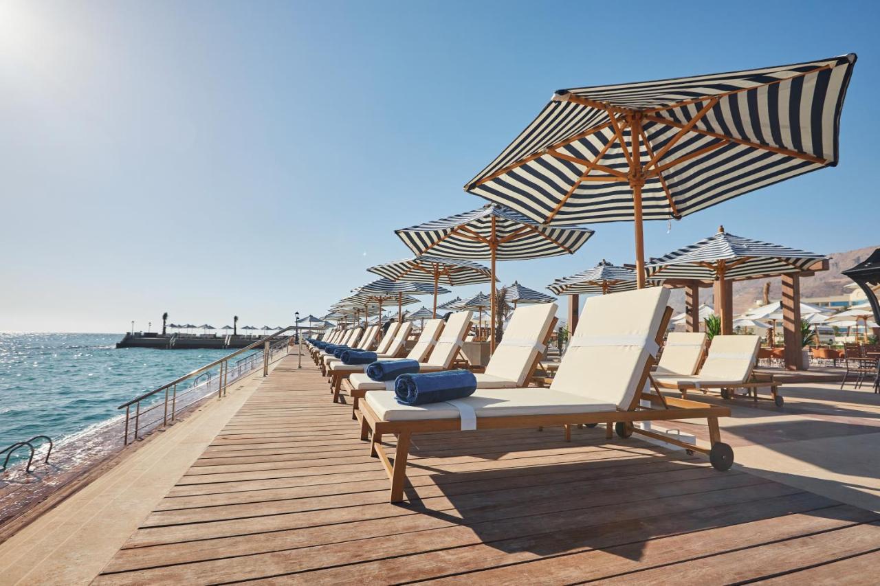 منتجع توليب الجلالة ماجستيك عين السخنة المظهر الخارجي الصورة The photo depicts a serene beachside setting featuring a row of sun loungers arranged on a wooden deck. Each lounger is equipped with a folded towel and shaded by striped umbrellas. In the background, the water sparkles under bright sunlight, and the