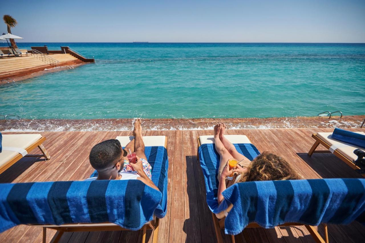 منتجع توليب الجلالة ماجستيك عين السخنة المظهر الخارجي الصورة The photo depicts two people relaxing on lounge chairs by the waterfront. They are positioned on a wooden deck with a view of the clear, turquoise water. Both individuals are reclining, enjoying the sun, and each has a drink in hand. The scene convey