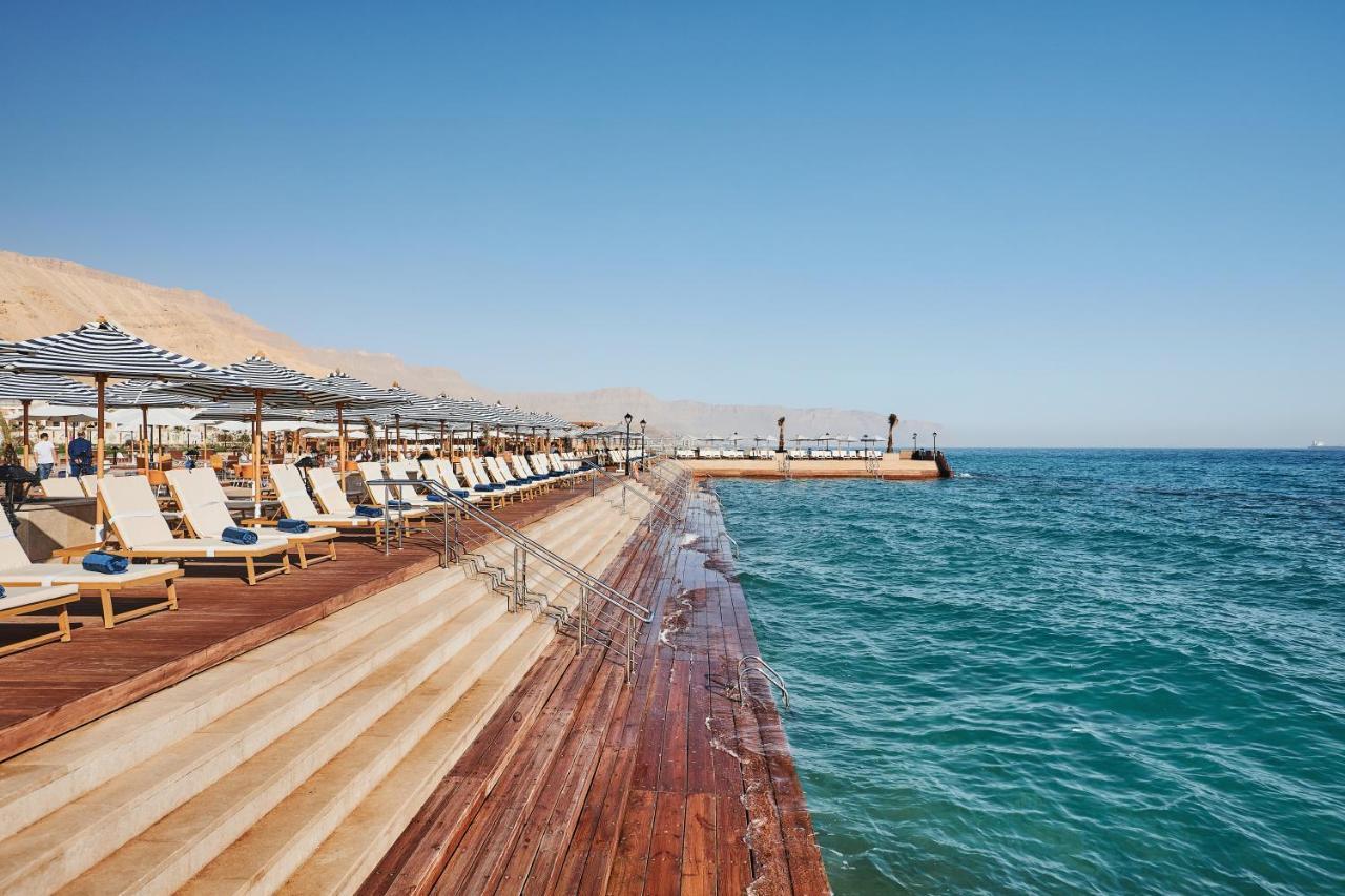منتجع توليب الجلالة ماجستيك عين السخنة المظهر الخارجي الصورة The photo shows a seaside area featuring a wooden deck that extends into the water. On the left side, there are several lounge chairs arranged neatly under shade structures, suggesting a relaxing atmosphere. The ocean is visible in the foreground, wi