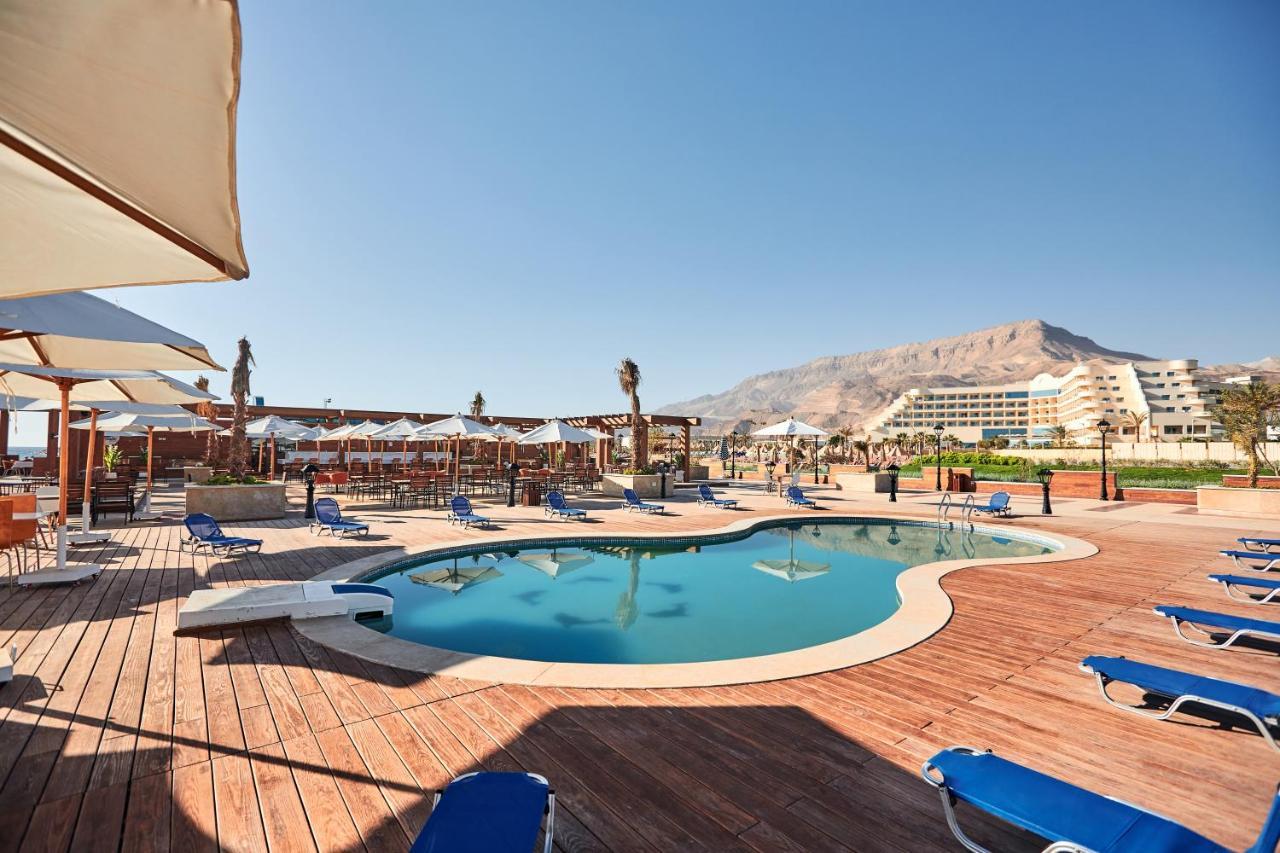 منتجع توليب الجلالة ماجستيك عين السخنة المظهر الخارجي الصورة The photo shows a swimming pool area surrounded by sun loungers and umbrellas. The pool has a unique, curvy shape and is situated on a wooden deck. In the background, there are several buildings, likely part of a resort or hotel, and a mountainous la