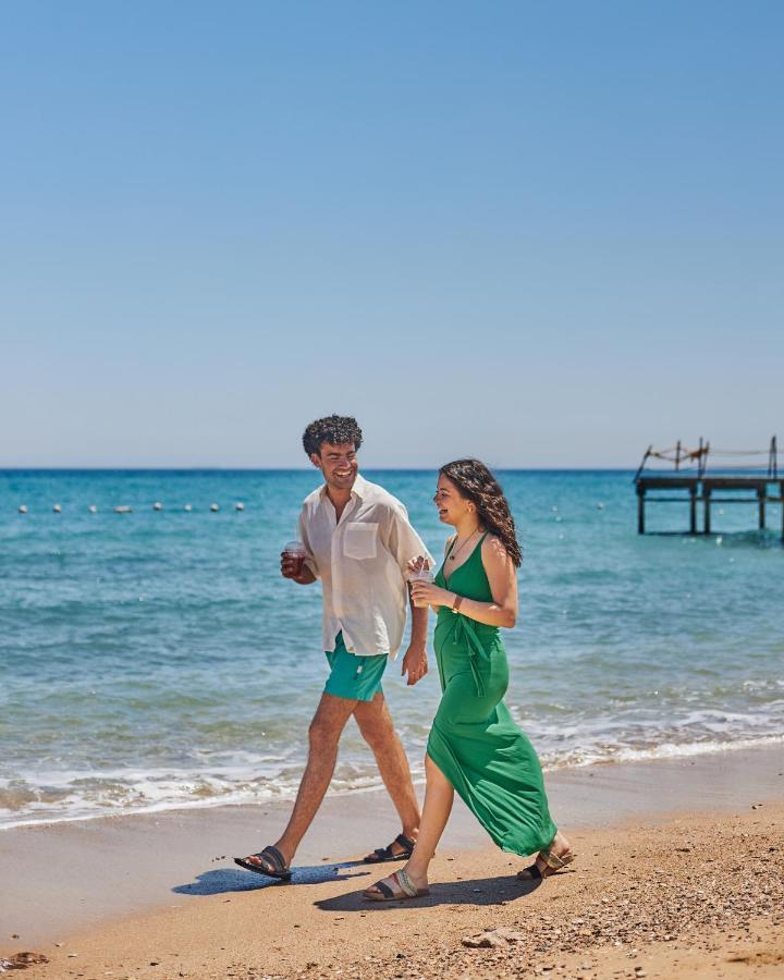 منتجع توليب الجلالة ماجستيك عين السخنة المظهر الخارجي الصورة The photo shows a couple walking along a beach. The man is wearing a white short-sleeved shirt and green shorts, while the woman is dressed in a flowing green dress. They both appear to be enjoying the pleasant weather, with the ocean in the backgrou
