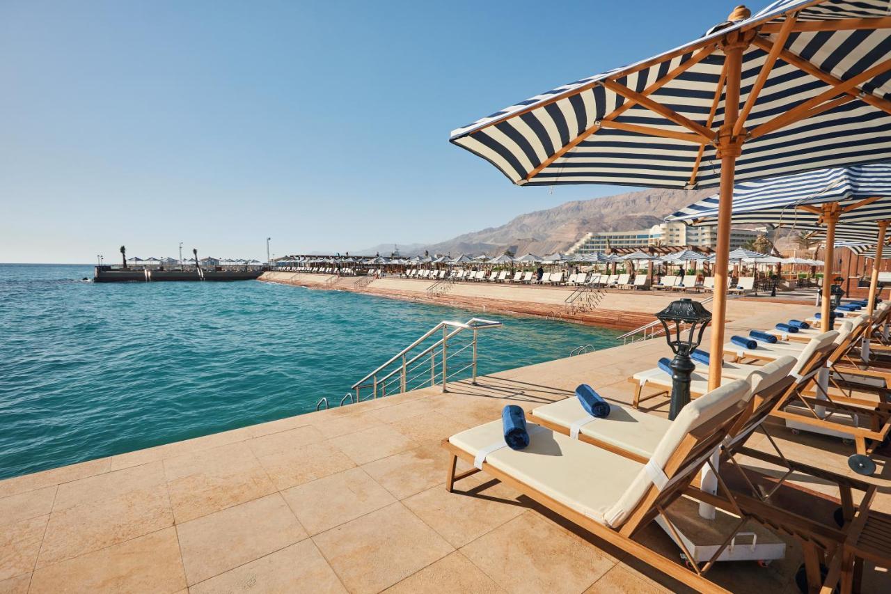 منتجع توليب الجلالة ماجستيك عين السخنة المظهر الخارجي الصورة The photo shows a scenic beachside area with clear blue water. In the foreground, there are several sun loungers set up on a tiled surface, with blue towels neatly rolled on each one. The loungers are shaded by striped umbrellas. In the distance, a w