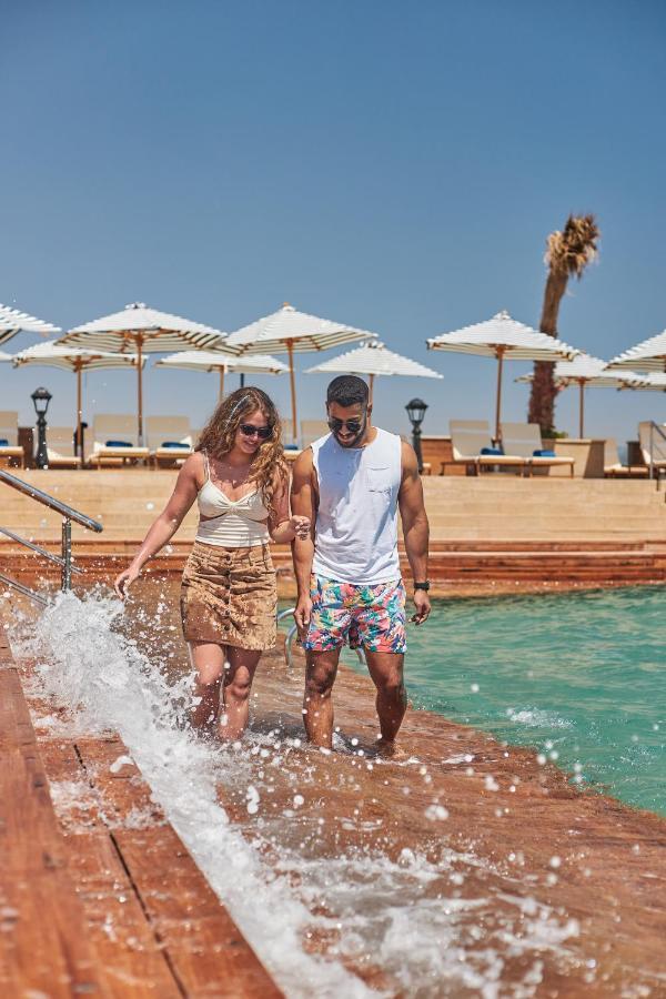 منتجع توليب الجلالة ماجستيك عين السخنة المظهر الخارجي الصورة The photo depicts a couple walking along a poolside area. They are enjoying a sunny day near a swimming pool, with splashes of water around them. The woman is wearing a light-colored top and a patterned skirt, while the man is dressed in a sleeveless