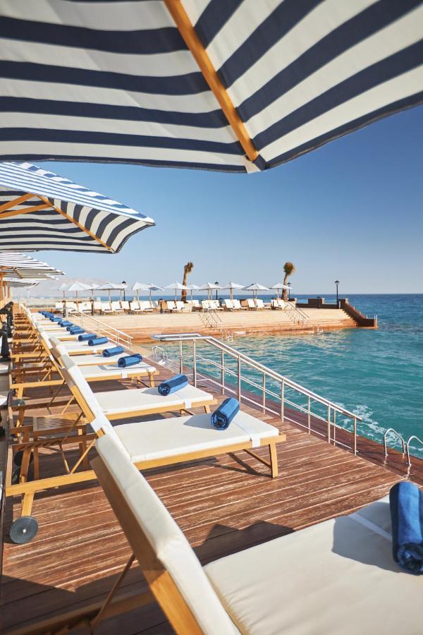منتجع توليب الجلالة ماجستيك عين السخنة المظهر الخارجي الصورة The photo depicts a serene outdoor area by the waterfront. There are several lounge chairs arranged in a row, each equipped with blue towels. Large, striped umbrellas provide shade over some of the chairs. In the background, there is a wooden deck th