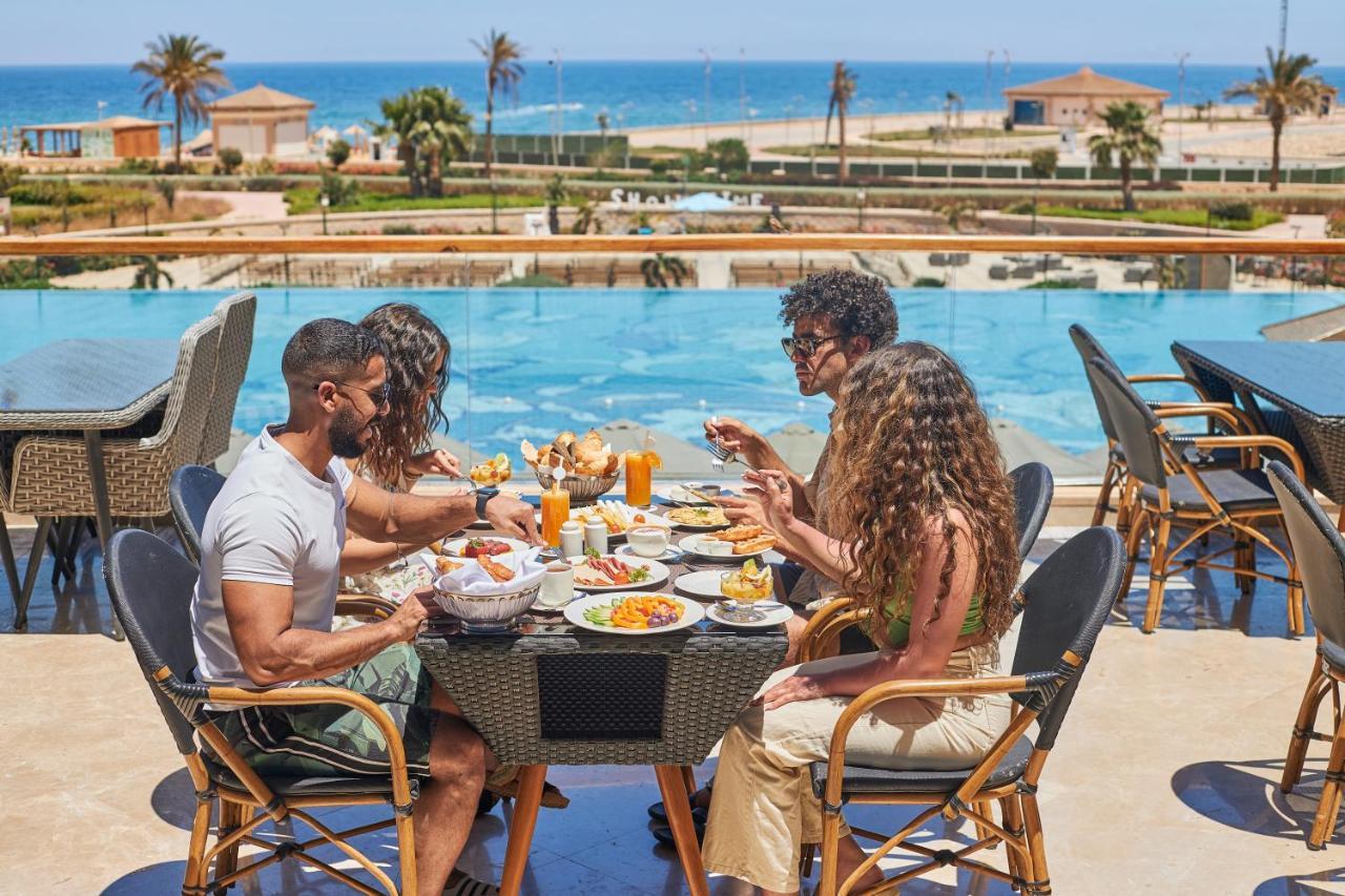 منتجع توليب الجلالة ماجستيك عين السخنة المظهر الخارجي الصورة The photo shows a group of five people enjoying a meal together outdoors, likely at a restaurant or cafe. They are seated at a table with a beautiful view of a swimming pool and the sea in the background. The table is filled with various dishes, and 