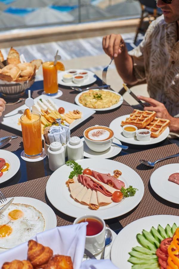 منتجع توليب الجلالة ماجستيك عين السخنة المظهر الخارجي الصورة The photo shows a breakfast spread laid out on a table. There are several plates with a variety of food items, including:

1. **Sliced Cold Cuts**: A plate featuring various cold meats, accompanied by garnishes like cherry tomatoes and lettuce.
2. **