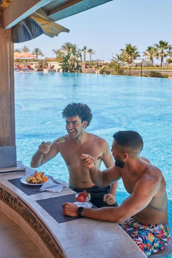 منتجع توليب الجلالة ماجستيك عين السخنة المظهر الخارجي الصورة The photo features two young men enjoying a meal together. They are sitting at a poolside table with a clear blue swimming pool in the background. The men are shirtless, suggesting a casual, relaxed atmosphere, likely in a warm environment. One man i