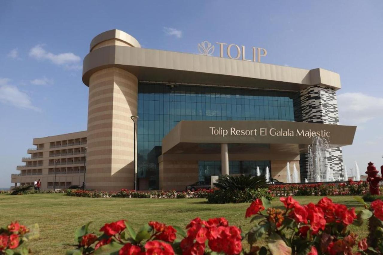 منتجع توليب الجلالة ماجستيك عين السخنة المظهر الخارجي الصورة The photo shows a modern hotel building named "Tolip Resort El Galala Majestic." The structure features a distinctive architectural design with large glass windows and a prominent entrance. Surrounding the building are well-maintained lawns and vibra