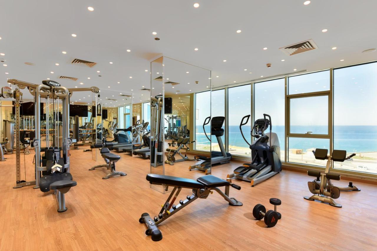 منتجع توليب الجلالة ماجستيك عين السخنة المظهر الخارجي الصورة The photo shows a modern gym interior with a variety of fitness equipment. There are cardio machines like treadmills and ellipticals, along with strength training equipment such as free weights and benches. The gym features large windows that offer a
