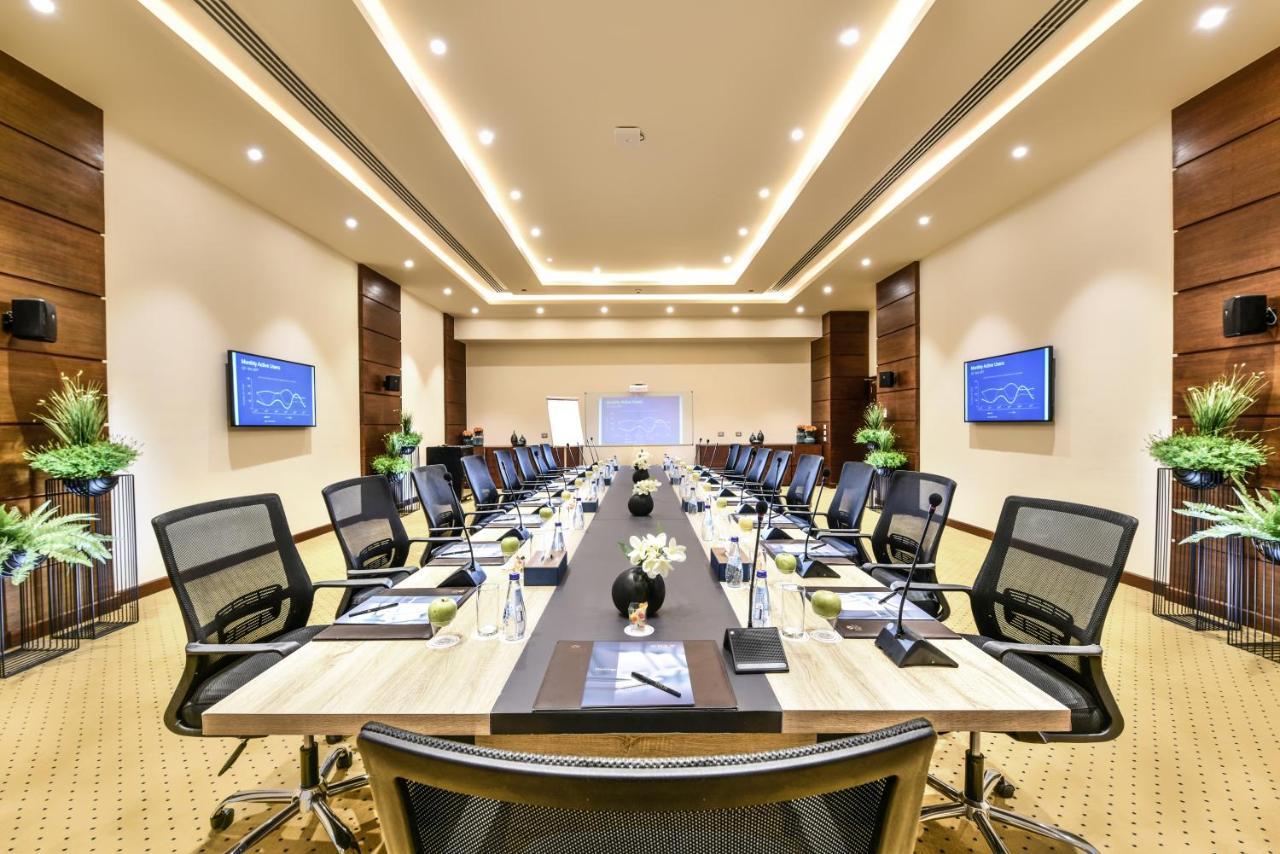 منتجع توليب الجلالة ماجستيك عين السخنة المظهر الخارجي الصورة The photo shows a modern conference room designed for meetings. It features a long rectangular table at the center, surrounded by ergonomic black chairs. The room is well-lit with ceiling lights and has a neutral color scheme. There are two large scr