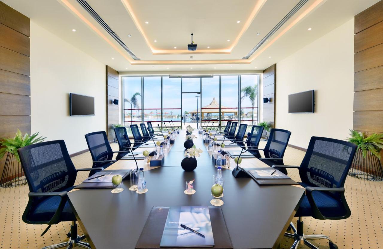 منتجع توليب الجلالة ماجستيك عين السخنة المظهر الخارجي الصورة The image shows a modern conference room with a large, dark rectangular table surrounded by ergonomic black chairs. The room features large windows that allow natural light to enter and provide a view of the outdoors, including some greenery. On the 