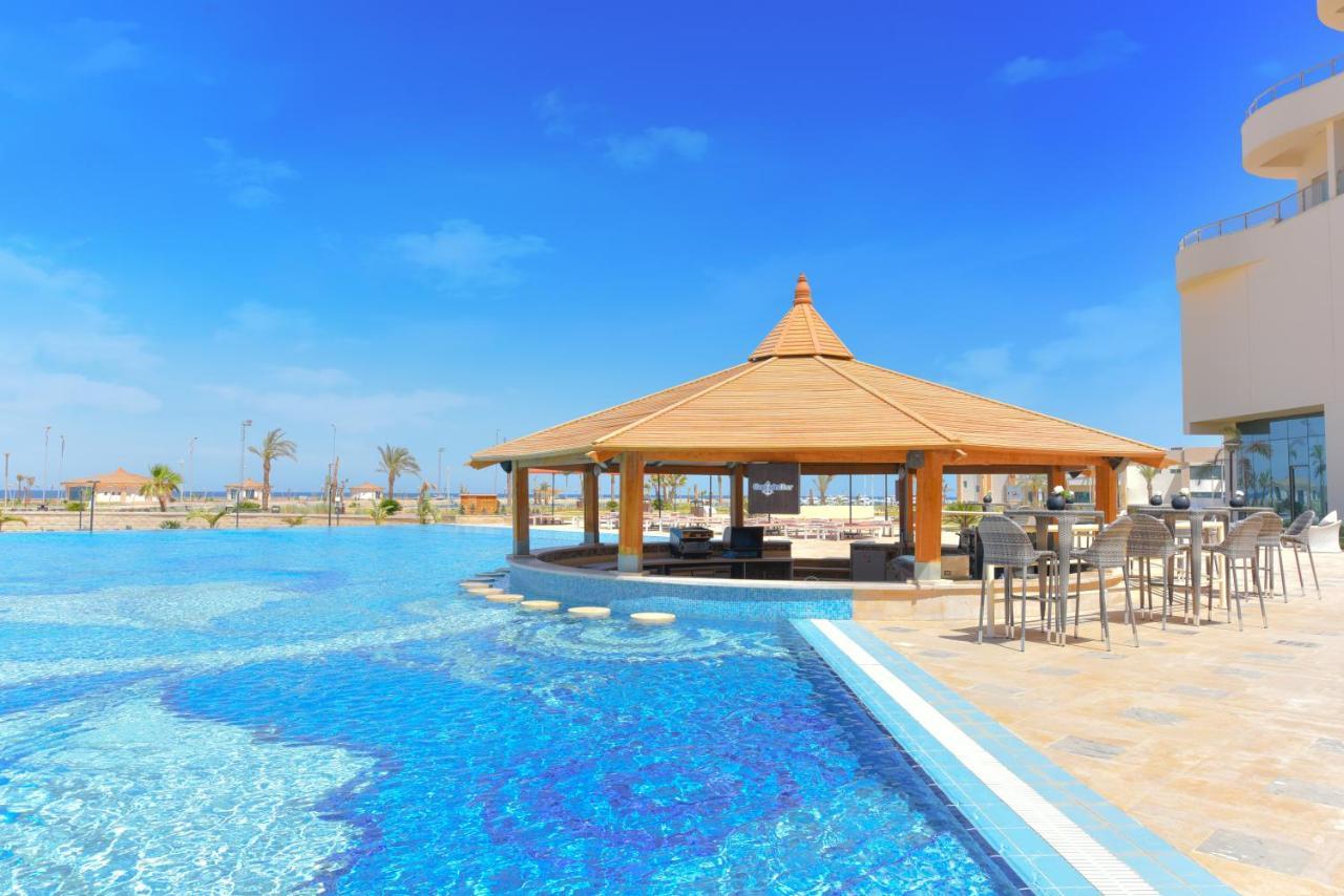 منتجع توليب الجلالة ماجستيك عين السخنة المظهر الخارجي الصورة The photo shows a beautiful outdoor setting featuring a large swimming pool with clear blue water. In the center, there is a thatched-roof gazebo, which serves as a shaded seating area. Surrounding the gazebo are lounge chairs and a bar area with hig