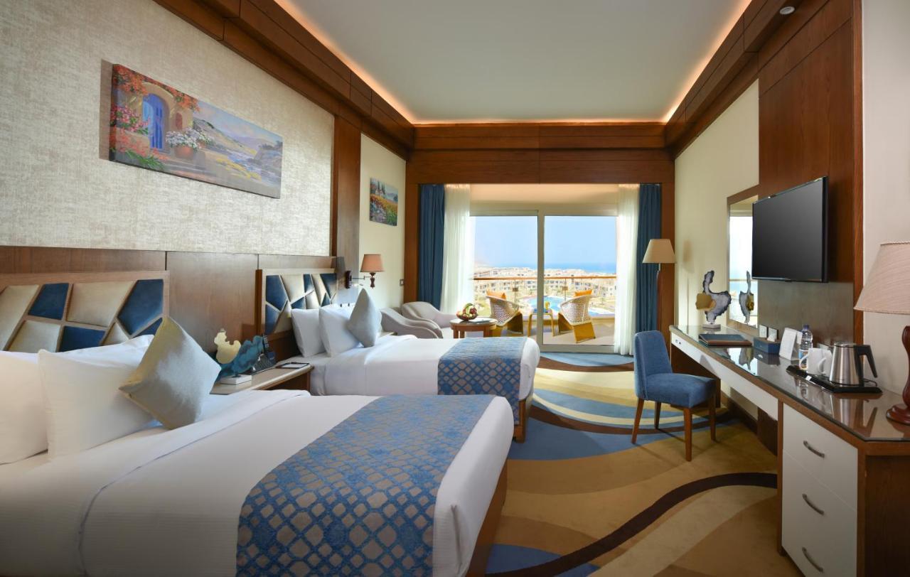 منتجع توليب الجلالة ماجستيك عين السخنة المظهر الخارجي الصورة The photo shows a well-decorated hotel room featuring two beds with decorative pillows. The room has wooden accents and modern furnishings, including a desk, a television, and lamps. Large windows flood the space with natural light, offering a view o