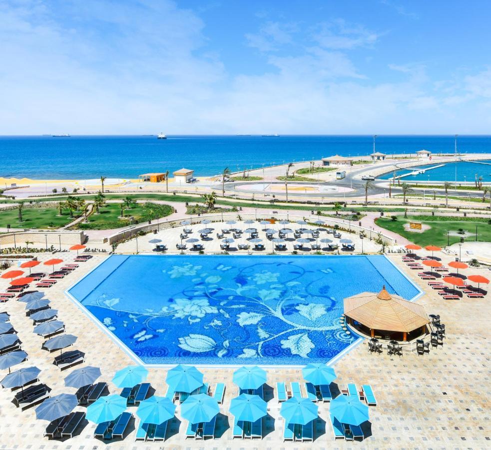 منتجع توليب الجلالة ماجستيك عين السخنة المظهر الخارجي الصورة The photo features a large swimming pool with a decorative design on its surface, surrounded by sun loungers and umbrellas. In the background, there is a view of the sea, with a clear blue sky and a few scattered clouds. The area is well-maintained, 