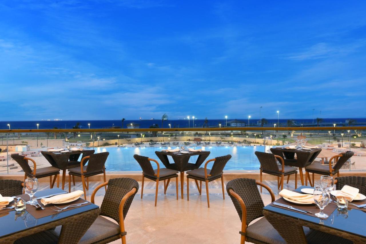 منتجع توليب الجلالة ماجستيك عين السخنة المظهر الخارجي الصورة The photo shows an outdoor dining area with several neatly arranged tables and chairs. The tables are elegantly set with cutlery and plates, suggesting a fine dining experience. In the background, there is a large swimming pool that reflects the blue