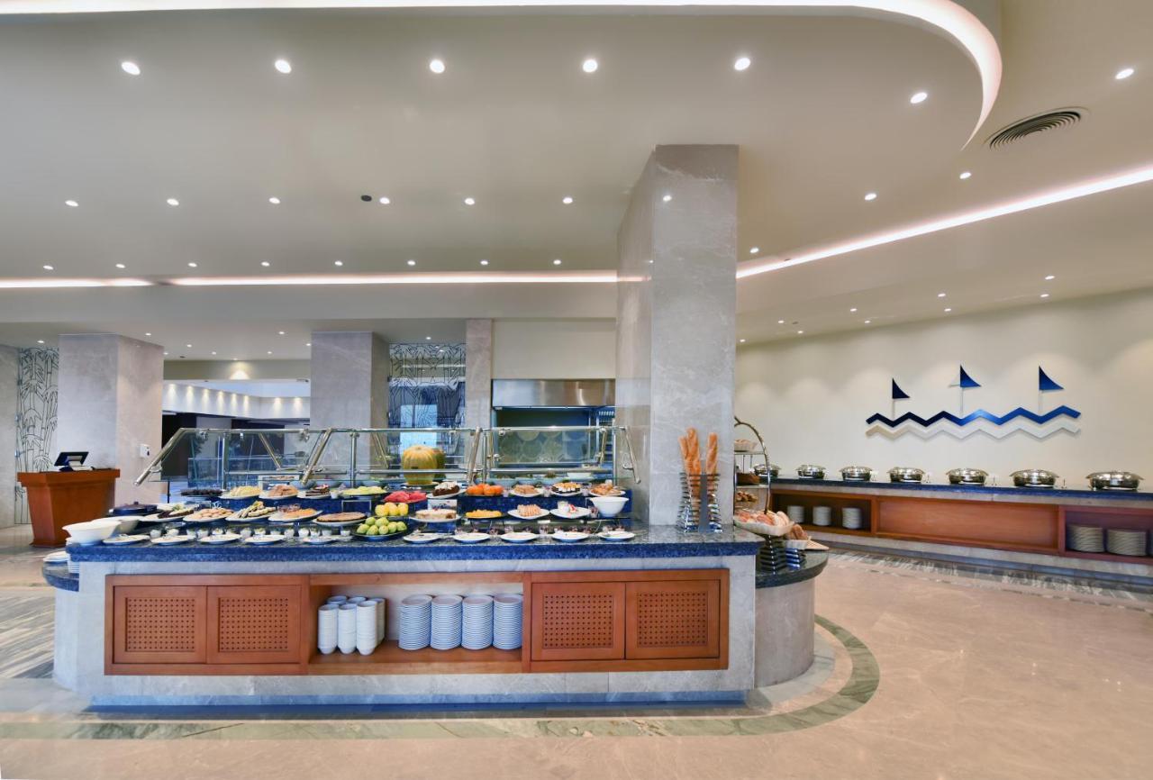 منتجع توليب الجلالة ماجستيك عين السخنة المظهر الخارجي الصورة The image shows a buffet-style dining area in a restaurant or hotel. There are several food stations with a variety of dishes displayed, including fruits, salads, and other food items. The layout features a large, central island with a wooden base an