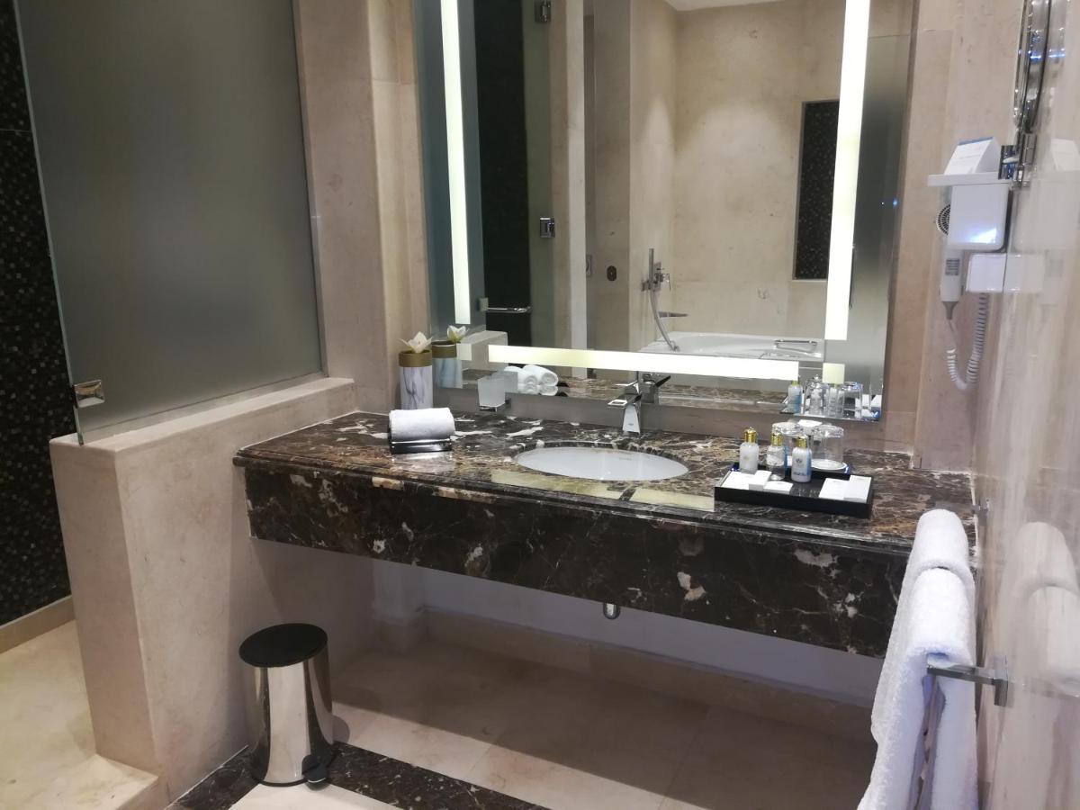 منتجع توليب الجلالة ماجستيك عين السخنة المظهر الخارجي الصورة The photo shows a modern bathroom with elegant design features. There is a large vanity made of dark marble with a sink and a set of toiletries neatly arranged on top. A mirror with built-in lighting is mounted above the sink. To the side, there is a