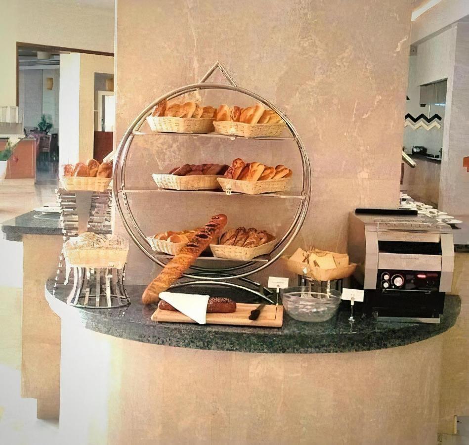 منتجع توليب الجلالة ماجستيك عين السخنة المظهر الخارجي الصورة The photo shows a display of various types of bread and pastries arranged on a countertop. There are several baskets stacked on a circular stand, filled with different baked goods like croissants and rolls. A long baguette is placed horizontally on a