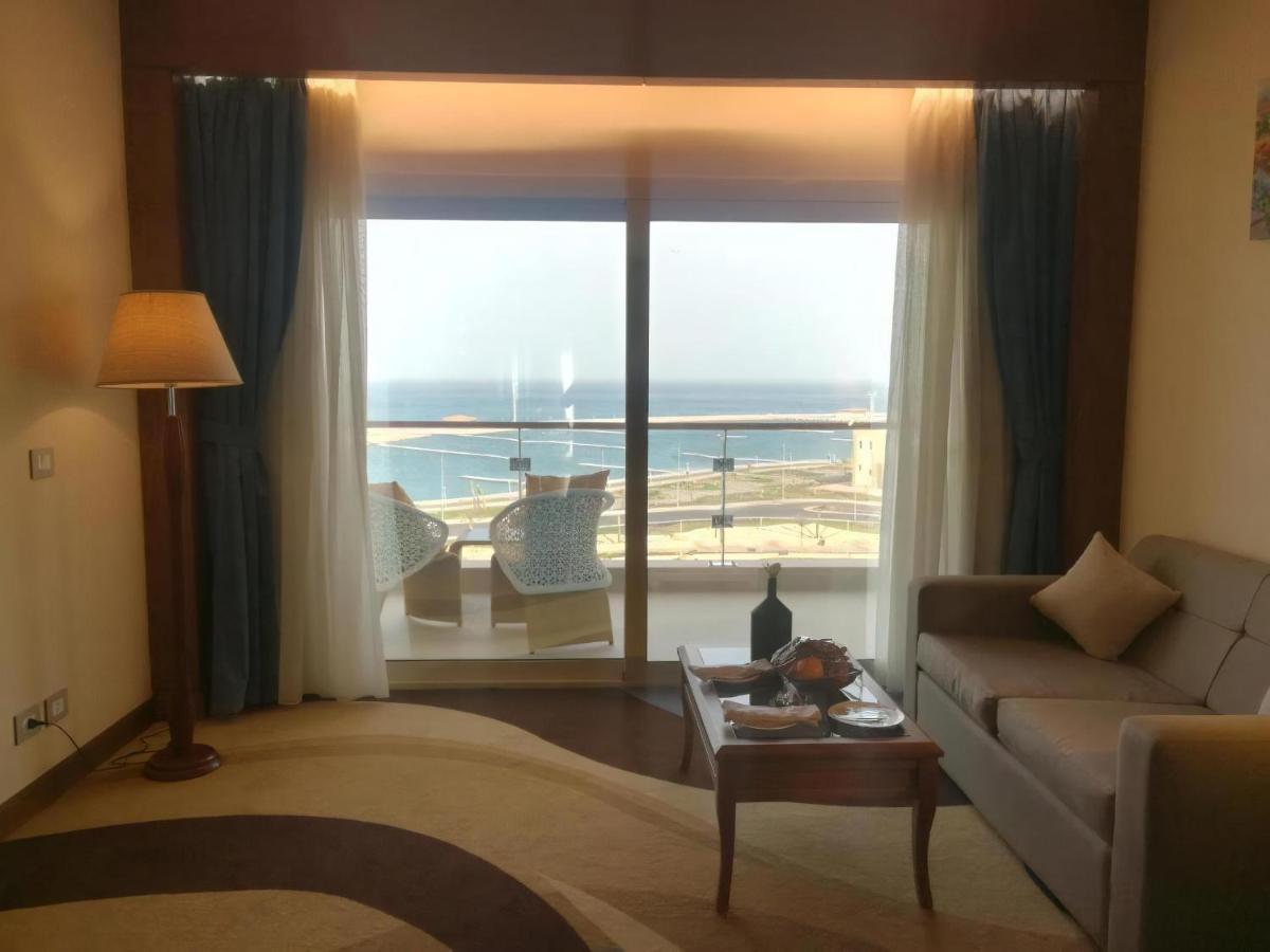 منتجع توليب الجلالة ماجستيك عين السخنة المظهر الخارجي الصورة The photo shows a stylish interior of a room with a large window overlooking the sea. The window features sheer and thick curtains and is framed by a balcony with two chairs visible outside. Inside, there is a comfortable-looking sofa, a coffee table