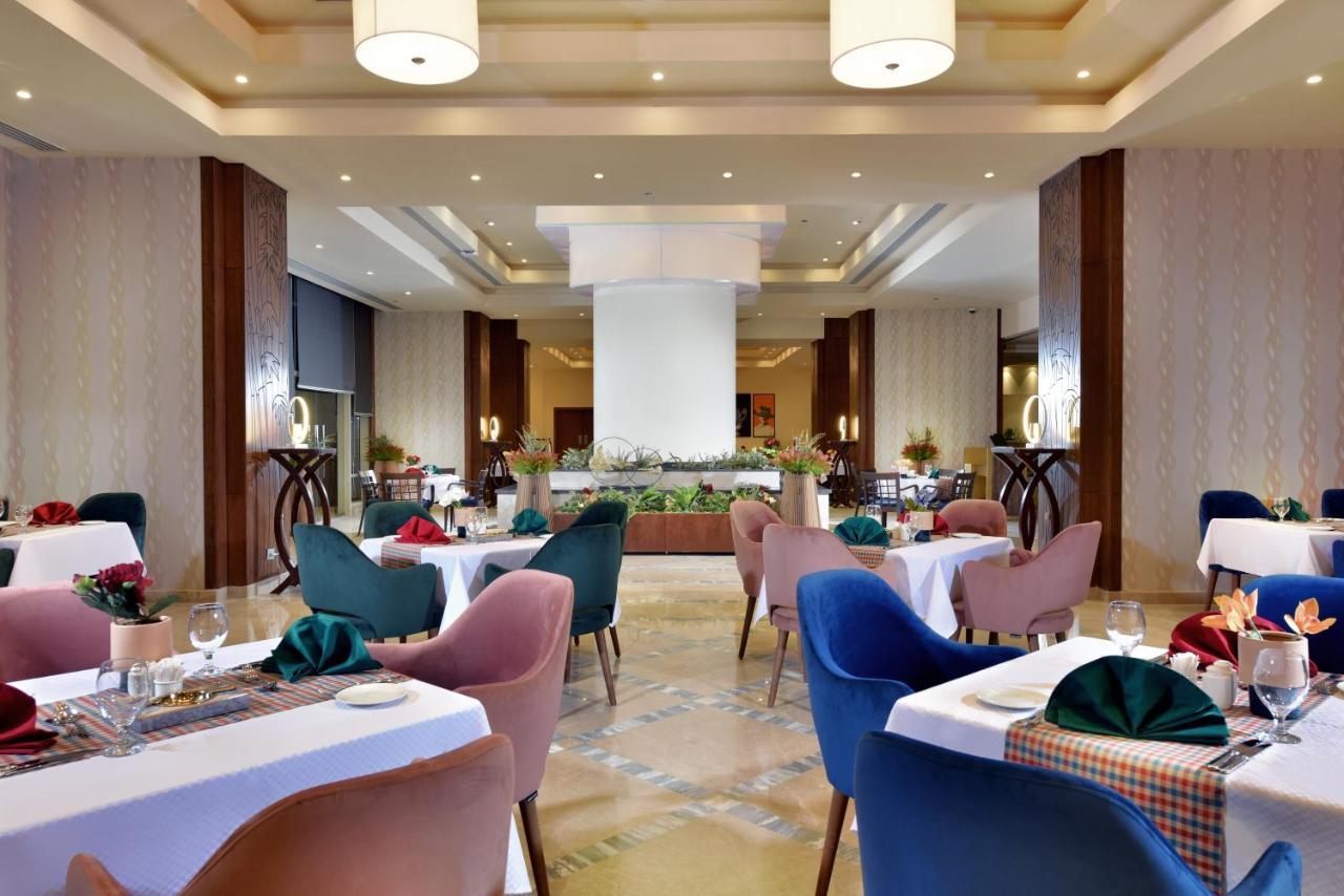 منتجع توليب الجلالة ماجستيك عين السخنة المظهر الخارجي الصورة The photo shows a spacious and elegantly designed restaurant interior. The layout includes several round tables dressed in white tablecloths, each set with decorative plates, cutlery, and colorful napkins. The chairs are upholstered in a variety of c
