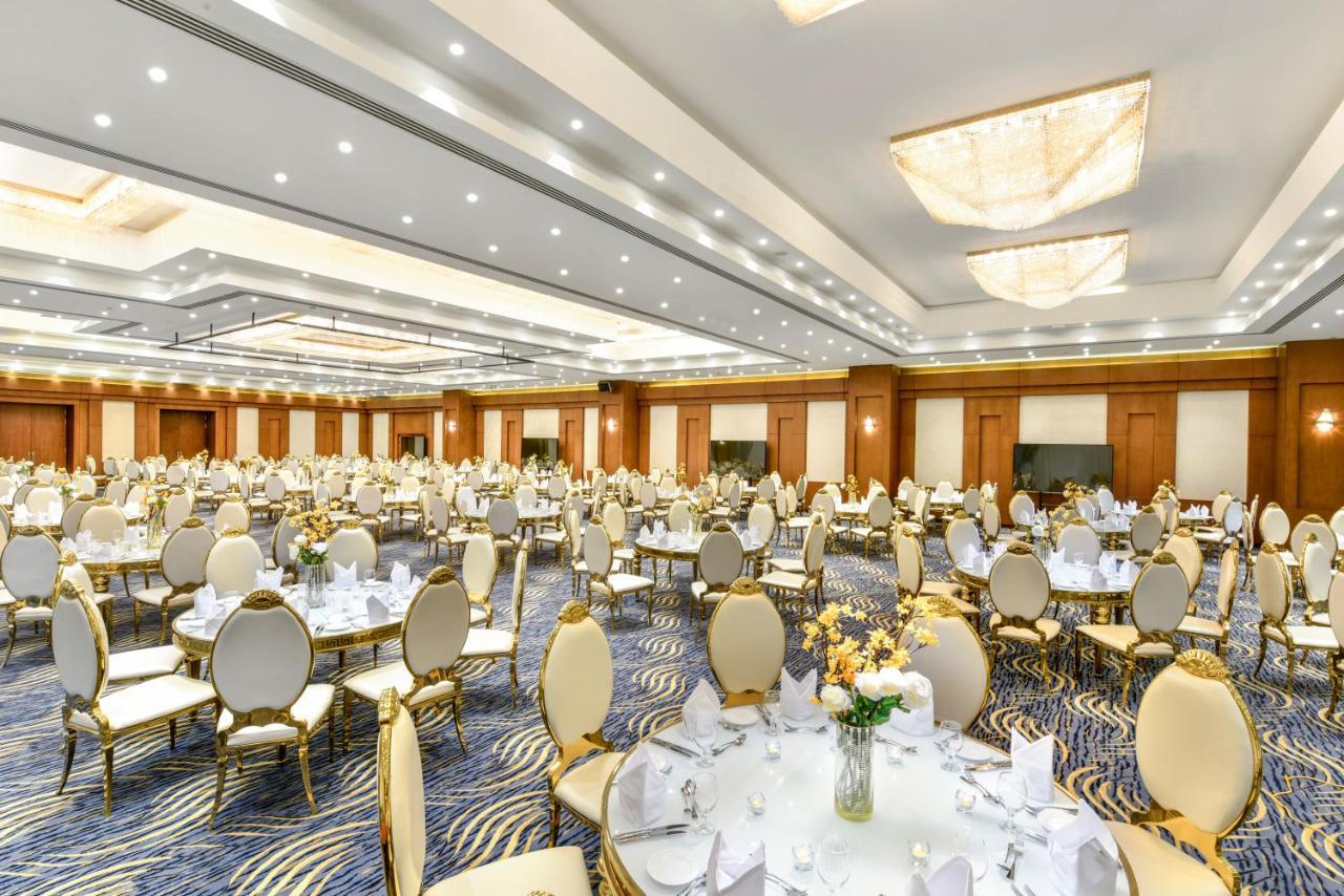 منتجع توليب الجلالة ماجستيك عين السخنة المظهر الخارجي الصورة The photo shows a spacious and elegantly decorated banquet hall. The room features numerous round dining tables, each set with white tablecloths and adorned with decorative centerpieces, likely flowers. The chairs surrounding the tables have a luxuri